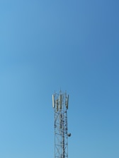 A tall communication tower stands against a clear blue sky. The structure is composed of a metal framework with several antennas and dishes attached to it.