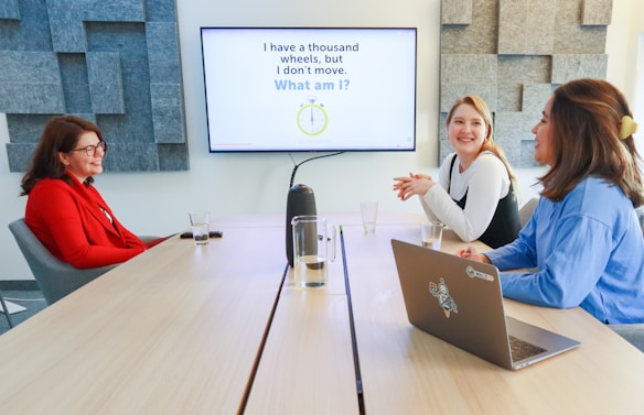 Three people are sitting around a conference table having a meeting. A screen on the wall displays a question written in large letters. The room has modern acoustic panels on the walls and a sleek, uncluttered appearance. Two people are sitting on one side of the table with a laptop in front of them, while the third person is sitting on the opposite side, engaged in conversation.