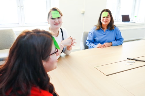 Three individuals are sitting around a wooden conference table. They are engaged in a game where each person has a green sticky note on their forehead with names written on them. The room is brightly lit with natural light coming through large windows. A laptop, marker, and some round tokens are placed on the table.