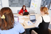 Three people are sitting around a conference table in a modern office. One person is wearing a red blazer and is speaking, while the other two listen and have open laptops in front of them. Papers with charts and graphs are scattered on the table. In the background, there are gray acoustic panels on the wall and a flip chart with colorful notes.
