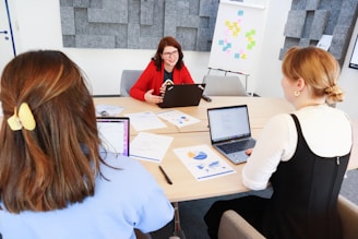 Three people are sitting around a conference table in a modern office. One person is wearing a red blazer and is speaking, while the other two listen and have open laptops in front of them. Papers with charts and graphs are scattered on the table. In the background, there are gray acoustic panels on the wall and a flip chart with colorful notes.