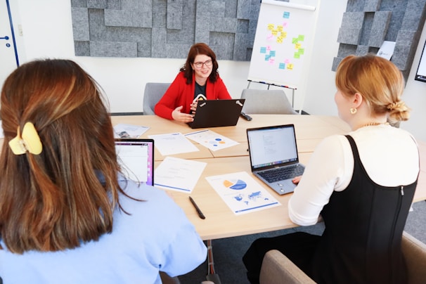 Three people are sitting around a conference table in a modern office. One person is wearing a red blazer and is speaking, while the other two listen and have open laptops in front of them. Papers with charts and graphs are scattered on the table. In the background, there are gray acoustic panels on the wall and a flip chart with colorful notes.