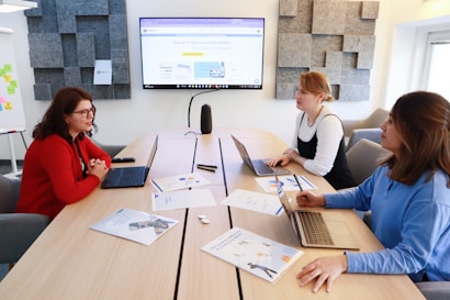 Three women are seated around a wooden table in a modern office setting, each with a laptop. A large screen on the wall displays a professional presentation. One woman wears a red sweater and glasses, another a white long-sleeved top with a black dress, and the third a blue shirt. Various documents and notepads are spread across the table, suggesting a meeting or collaborative work session. Acoustic panels are on the walls.