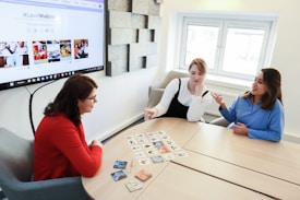 Three women are sitting around a wooden table, engaged in a discussion while looking at cards laid out on the table. One woman is wearing a red sweater, another a white top with a black overdress, and the third a blue shirt. A large screen displaying a website with the hashtag #LoveWallsio is mounted on the wall behind them. The room has a bright, modern aesthetic with a window letting in natural light.
