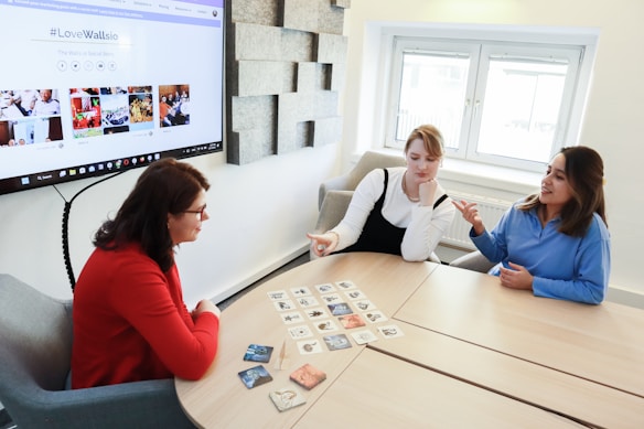 Three women are sitting around a wooden table, engaged in a discussion while looking at cards laid out on the table. One woman is wearing a red sweater, another a white top with a black overdress, and the third a blue shirt. A large screen displaying a website with the hashtag #LoveWallsio is mounted on the wall behind them. The room has a bright, modern aesthetic with a window letting in natural light.