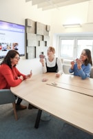 Three people are sitting around a light wooden table, engaged in a card game. The person on the left is wearing a red blazer, while the person in the middle is in a white shirt and black top. The person on the right is wearing a blue sweater. A computer screen on the left wall displays a webpage, and a textured gray art piece is mounted on the wall nearby. A window with white blinds is visible in the background.