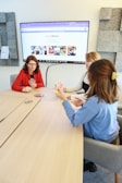 Three people are sitting around a light wooden table playing a card game. One person in a red top holds a hand of cards, while the others are also engaged with their cards. A large screen on the wall displays a website with the hashtag #LoveWallsio.