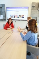 Three people are sitting around a light wooden table playing a card game. One person in a red top holds a hand of cards, while the others are also engaged with their cards. A large screen on the wall displays a website with the hashtag #LoveWallsio.
