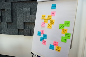 A flip chart with various colored sticky notes attached, containing words and concepts like 'people 1st', 'respect', 'diversity', 'vision', and 'teamwork'. The notes are predominantly pink, yellow, green, and blue. The background features a wall with geometric, dark gray panels.
