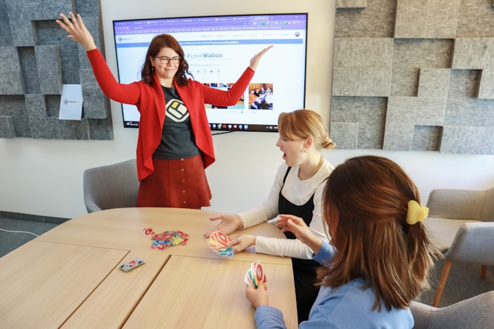 Four people are engaged in a lively game around a wooden table. One person stands with raised arms, while three others are seated, focusing on colorful cards in their hands. There is a computer monitor in the background displaying a webpage, and the room features a modern design with gray acoustic panels on the walls.