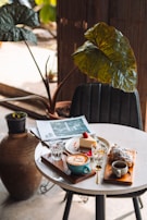 A cozy cafe table setup featuring a cappuccino in a blue mug, a slice of cheesecake garnished with a strawberry, a croissant dusted with powdered sugar, and two glasses of water. A folded magazine lies next to the plate, and the table is positioned near large potted plants, adding to the tranquil and inviting atmosphere.