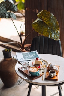 A cozy cafe table setup featuring a cappuccino in a blue mug, a slice of cheesecake garnished with a strawberry, a croissant dusted with powdered sugar, and two glasses of water. A folded magazine lies next to the plate, and the table is positioned near large potted plants, adding to the tranquil and inviting atmosphere.