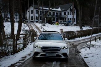 A white car is parked on a snow-covered driveway leading to a two-story house. The house is surrounded by trees, and there is snow on the ground and rooftops. The driveway is flanked by fences and bushes.