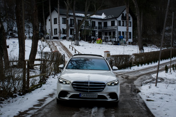 A white car is parked on a snow-covered driveway leading to a two-story house. The house is surrounded by trees, and there is snow on the ground and rooftops. The driveway is flanked by fences and bushes.