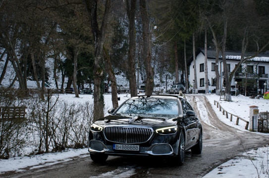 A luxury car is parked on a snow-covered road flanked by trees and a modern house in the background. The scene has a wintry setting with bare trees and patches of snow, creating a contrast with the sleek, dark car.