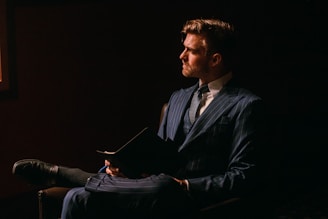 A man in a dark, elegant setting wearing a pinstripe navy suit. He sits in a chair with his legs crossed, holding an open book or folder. The lighting creates a dramatic contrast, highlighting his facial features while casting shadows around him.