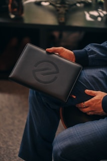 A person holding a black leather clutch with a debossed circular logo, sitting on a chair. The person is wearing a blue checkered suit and has a ring on their finger. The background features a dark table with blurred objects.