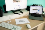 A modern workspace featuring a large monitor and an open laptop, both displaying content related to social media. A piece of paper titled 'Content Strategy' lies on the desk, accompanied by a keyboard, mouse, and some small tech accessories. The background is a teal color, adding a vibrant touch to the setup.