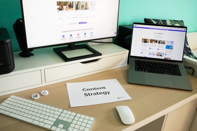 A modern workspace featuring a large monitor and an open laptop, both displaying content related to social media. A piece of paper titled 'Content Strategy' lies on the desk, accompanied by a keyboard, mouse, and some small tech accessories. The background is a teal color, adding a vibrant touch to the setup.