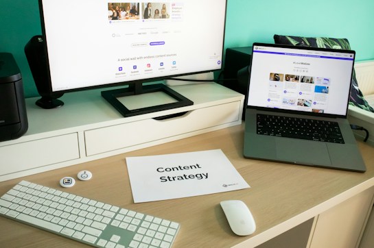 A modern workspace featuring a large monitor and an open laptop, both displaying content related to social media. A piece of paper titled 'Content Strategy' lies on the desk, accompanied by a keyboard, mouse, and some small tech accessories. The background is a teal color, adding a vibrant touch to the setup.