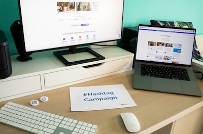 A modern workspace setup featuring a large monitor and a laptop displaying web pages. The desk has a wireless keyboard, a mouse, and a card labeled '#Hashtag Campaign'. The space is clean and organized with a focus on digital content.