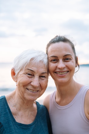 Two people smiling closely together, appearing happy and relaxed. The older person has short white hair and is wearing a blue top, while the younger person has pulled-back brown hair and is wearing a sleeveless light-colored top. The background features a serene seascape with a soft, cloudy sky.