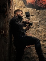 A person is holding a professional camera while standing against a stone wall. The scene is dimly lit, emphasizing the textures of the wall and the equipment. In the background, there is a glimpse of a sunset or sunrise with silhouettes of trees.