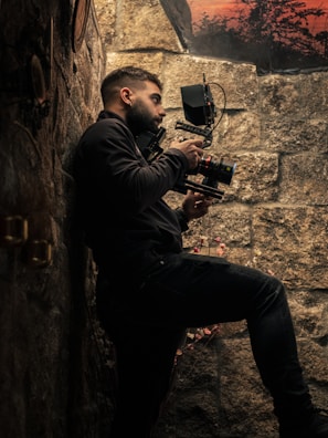 A person is holding a professional camera while standing against a stone wall. The scene is dimly lit, emphasizing the textures of the wall and the equipment. In the background, there is a glimpse of a sunset or sunrise with silhouettes of trees.