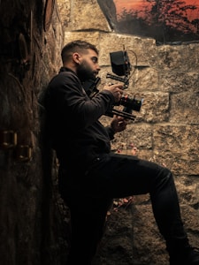 A person is holding a professional camera while standing against a stone wall. The scene is dimly lit, emphasizing the textures of the wall and the equipment. In the background, there is a glimpse of a sunset or sunrise with silhouettes of trees.