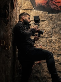 A person is holding a professional camera while standing against a stone wall. The scene is dimly lit, emphasizing the textures of the wall and the equipment. In the background, there is a glimpse of a sunset or sunrise with silhouettes of trees.