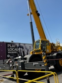 A large yellow construction crane is positioned in front of an urban billboard advertising 'Play Local Wilshire' with the Metro logo. The crane's metal sections and the machinery are prominent, and caution barriers surround the area.