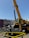 A large yellow construction crane is positioned in front of an urban billboard advertising 'Play Local Wilshire' with the Metro logo. The crane's metal sections and the machinery are prominent, and caution barriers surround the area.