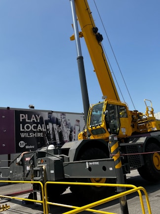 A large yellow construction crane is positioned in front of an urban billboard advertising 'Play Local Wilshire' with the Metro logo. The crane's metal sections and the machinery are prominent, and caution barriers surround the area.