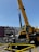 A large yellow construction crane is positioned in front of an urban billboard advertising 'Play Local Wilshire' with the Metro logo. The crane's metal sections and the machinery are prominent, and caution barriers surround the area.