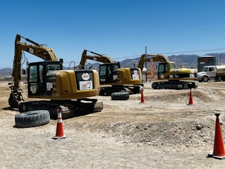 Several yellow excavators are lined up on a construction site with dirt and gravel on the ground. Bright orange traffic cones are placed around the area. In the background, a clear blue sky is visible above distant mountains.