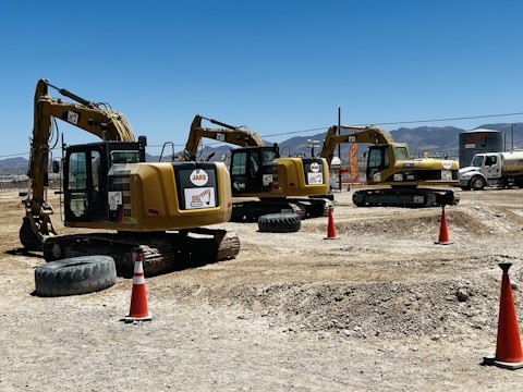 Several yellow excavators are lined up on a construction site with dirt and gravel on the ground. Bright orange traffic cones are placed around the area. In the background, a clear blue sky is visible above distant mountains.