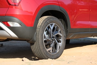 A red car is parked on sandy terrain, displaying its rear side and wheel prominently. The vehicle features sporty design elements, including a stylish alloy wheel and a sleek tail light. The ground is sandy with some small rocks scattered around.