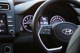 A modern car steering wheel with control buttons and a dashboard featuring a digital display and speedometer. The interior is sleek with visible media controls and air conditioning settings.