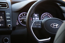 A modern car steering wheel with control buttons and a dashboard featuring a digital display and speedometer. The interior is sleek with visible media controls and air conditioning settings.