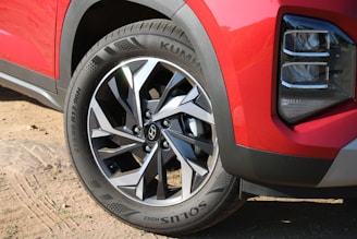 A close-up view of a car's front wheel featuring a stylish metal alloy rim with a modern design. The tire displays the brand name and specifications. The car's body is painted in a glossy red color, and part of a headlight is visible, suggesting a sporty appearance. The ground consists of sand and small patches of vegetation.