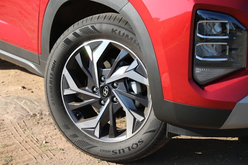 A close-up view of a car's front wheel featuring a stylish metal alloy rim with a modern design. The tire displays the brand name and specifications. The car's body is painted in a glossy red color, and part of a headlight is visible, suggesting a sporty appearance. The ground consists of sand and small patches of vegetation.