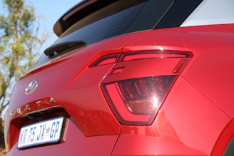 Close-up view of the back of a red Hyundai Creta, focusing on the rear brake light and license plate. The background shows some blurred greenery, indicating an outdoor setting.