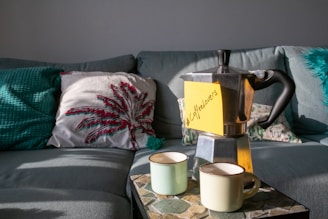 A cozy living room setup with a grey couch adorned with decorative pillows, including one with a red and green palm leaf design. On a stone-tiled table, there is a silver stovetop espresso maker with a yellow sticky note reading '#CoffeeLovers' attached to it. Two light-colored ceramic mugs are placed in front of the espresso maker.