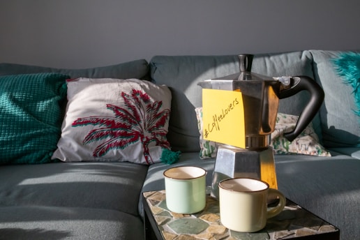 A cozy living room setup with a grey couch adorned with decorative pillows, including one with a red and green palm leaf design. On a stone-tiled table, there is a silver stovetop espresso maker with a yellow sticky note reading '#CoffeeLovers' attached to it. Two light-colored ceramic mugs are placed in front of the espresso maker.