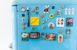 A blue refrigerator door is adorned with an assortment of colorful magnets, including circular and square shapes. Some of the magnets feature characters, animals, and various symbols. Among them is a yellow sticky note with the message '#Enjoy Cooking' written on it. The design styles range from playful to vintage, with images of moose, spaceships, and abstract patterns.