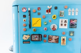 A blue refrigerator door is adorned with an assortment of colorful magnets, including circular and square shapes. Some of the magnets feature characters, animals, and various symbols. Among them is a yellow sticky note with the message '#Enjoy Cooking' written on it. The design styles range from playful to vintage, with images of moose, spaceships, and abstract patterns.
