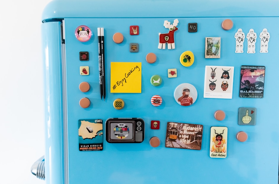 A blue refrigerator door is adorned with an assortment of colorful magnets, including circular and square shapes. Some of the magnets feature characters, animals, and various symbols. Among them is a yellow sticky note with the message '#Enjoy Cooking' written on it. The design styles range from playful to vintage, with images of moose, spaceships, and abstract patterns.