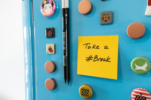 A light blue refrigerator door adorned with various colorful magnets and bottle caps. A bright yellow sticky note is attached with the message 'Take a #Break' written in black ink. Next to the note are two black pens held upright by a magnet.
