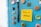 A light blue refrigerator door adorned with various colorful magnets and bottle caps. A bright yellow sticky note is attached with the message 'Take a #Break' written in black ink. Next to the note are two black pens held upright by a magnet.