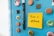 A light blue refrigerator door adorned with various colorful magnets and bottle caps. A bright yellow sticky note is attached with the message 'Take a #Break' written in black ink. Next to the note are two black pens held upright by a magnet.